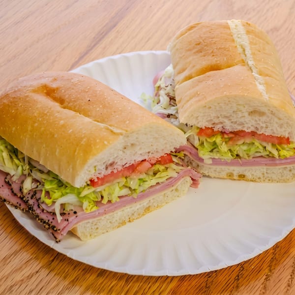 Two halves of a deli sub sandwich with ham, lettuce, and tomato on a white paper plate.