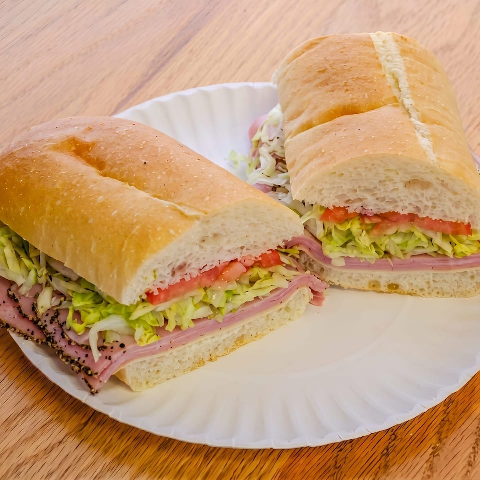 Two halves of a deli sub sandwich with ham, lettuce, and tomato on a white paper plate.