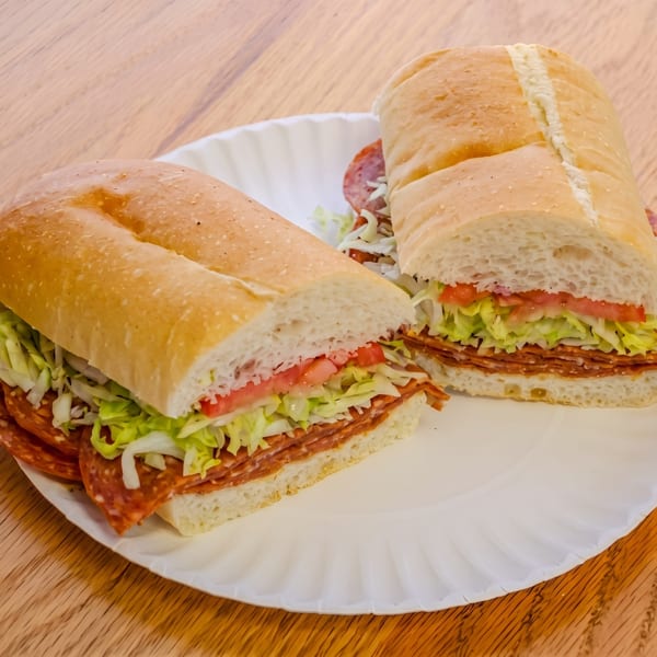 Two halves of a deli sub sandwich with salami, lettuce, and tomato on a white paper plate.