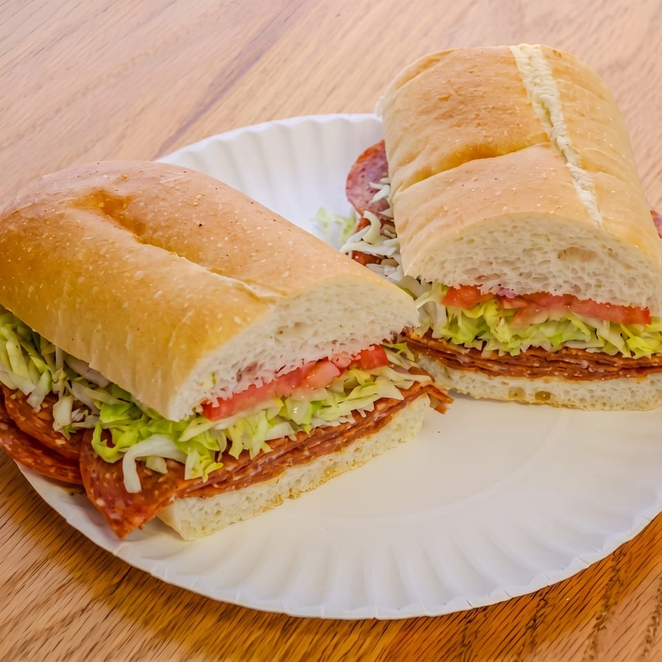 Two halves of a deli sub sandwich with salami, lettuce, and tomato on a white paper plate.